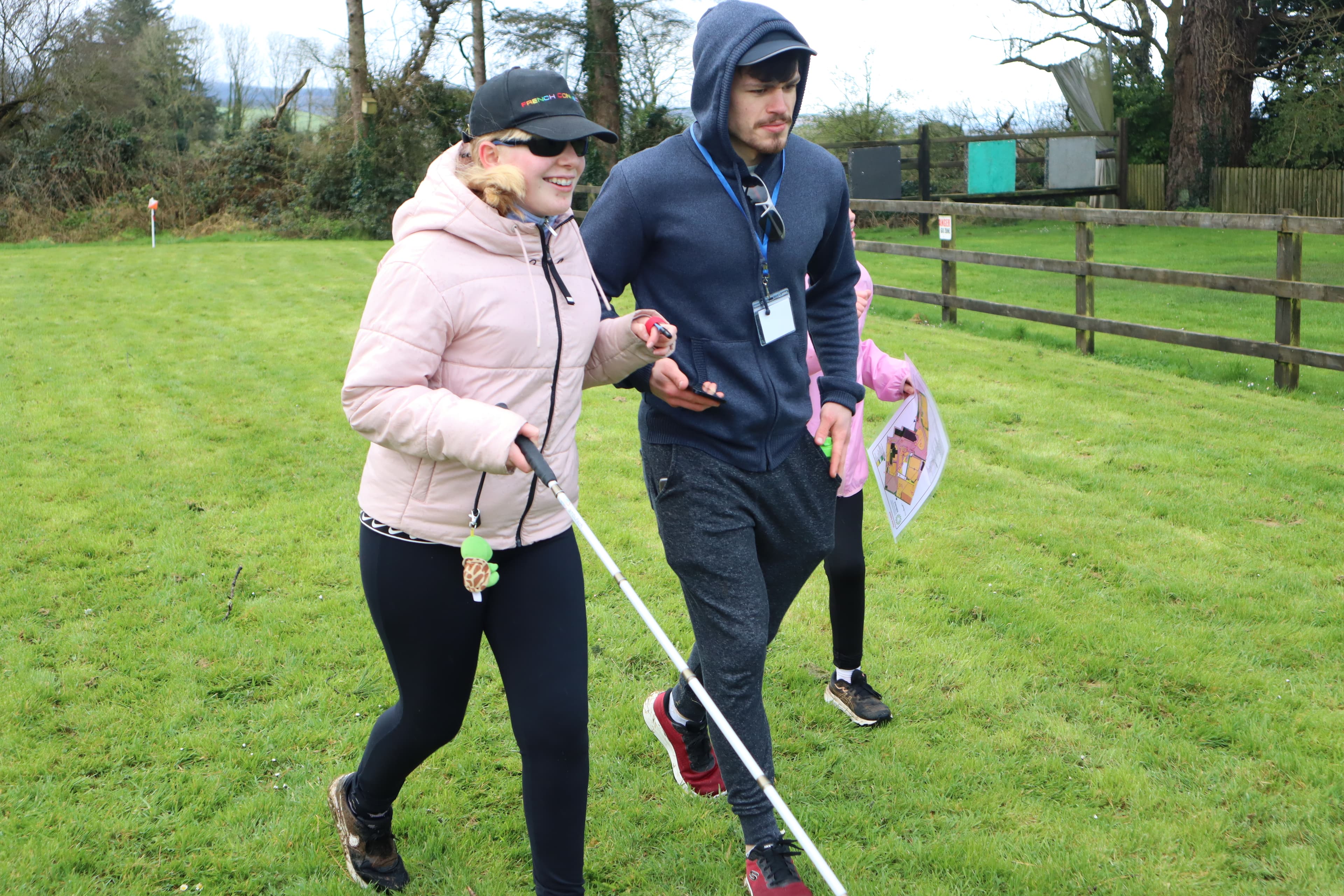 Three people walking across the feild, The person closest to the camera is using a white cane and the man next to her is holding a phone and assisting her movement. While a child follows them both from a slight distance.