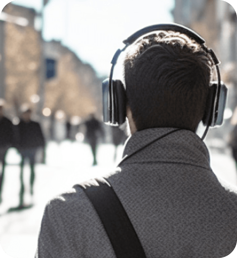 Image looking at the back of a man in a gray wool jacket with headphones on his head. As he is walking down a busy street using the Soundscape Community App.