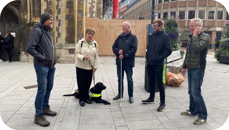 Picture of a group of people standing in an open space listening to Soundscape. One person has a guide dog, another person as a long cane.